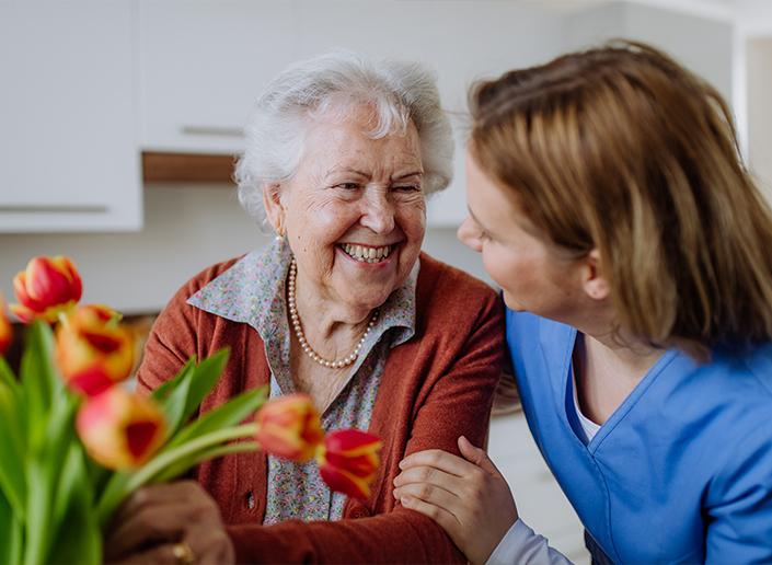 Senior care caregiver sharing flowers and warm conversation with smiling elderly woman at home